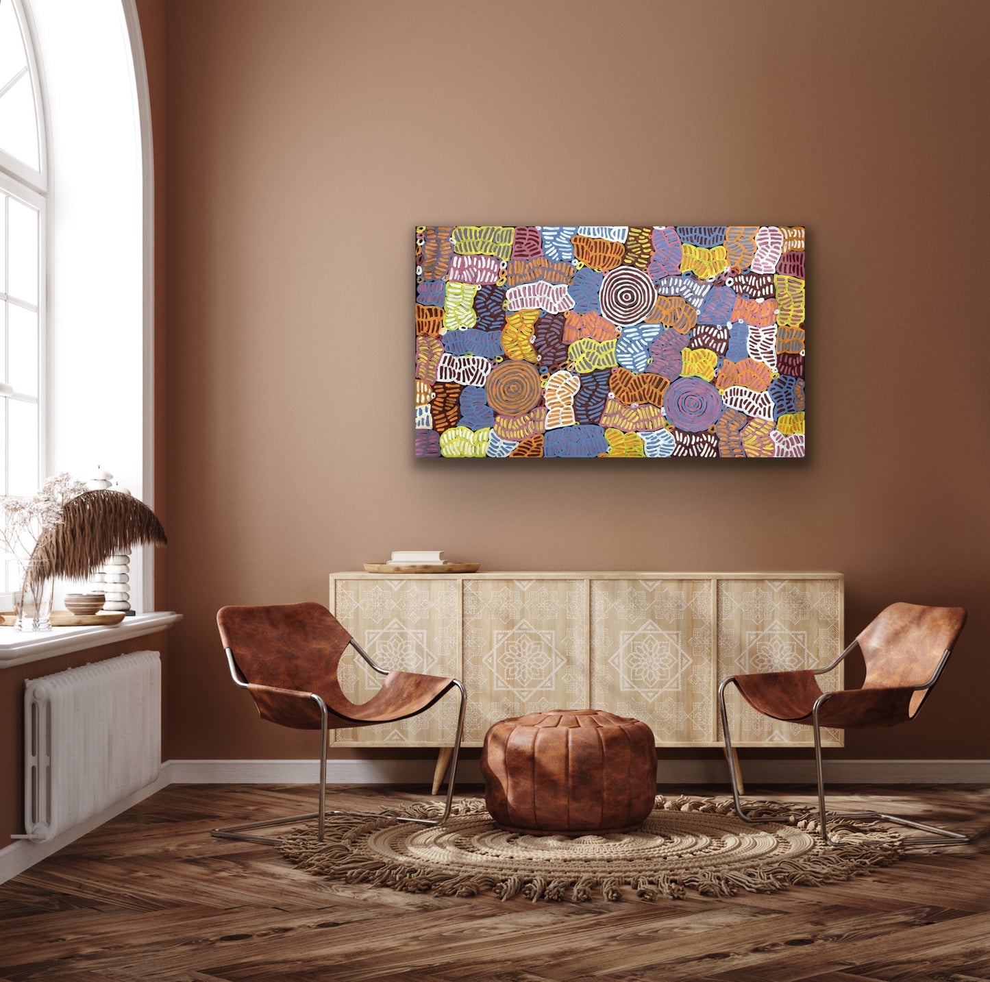 An awelye painting by Betty Club Mbitjana hanging on a wall above a wooden console and two brown leather chairs.