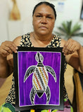 Artist Christine Burarrwanga holding her small painting depicting a sea turtle with traditional rarrk patterns on a purple background.