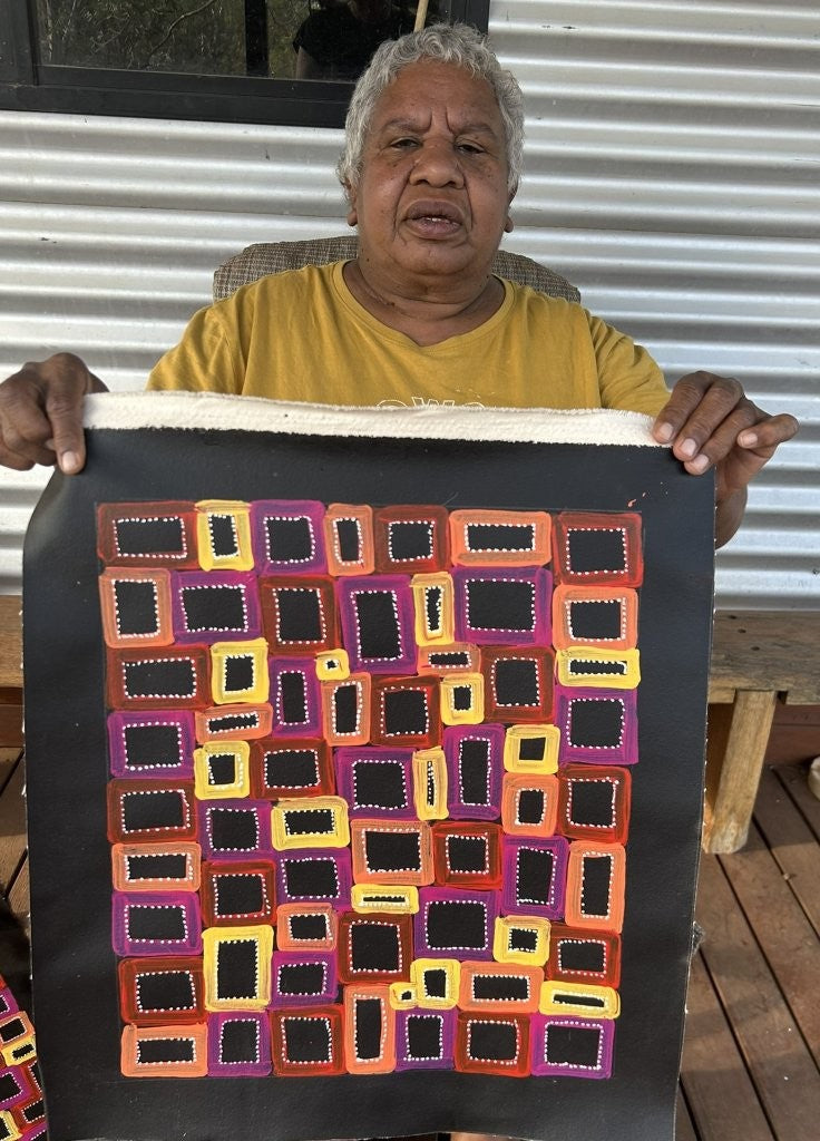 Artist Abie Loy Kemarre holding her small Awelye painting with colourful geometric patterns and dotting.