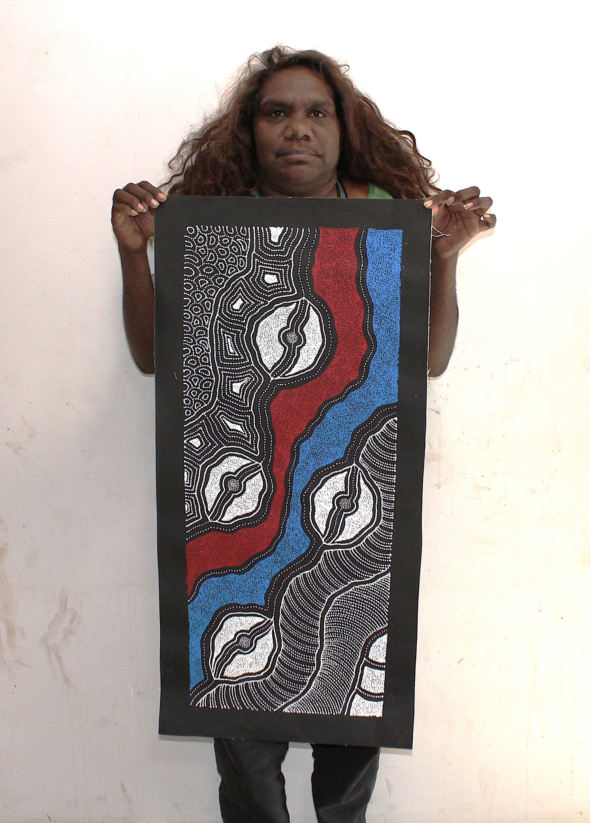 Delvine Pitjara holding her My Country painting with intricate dotted patterns in red, blue and white on black.