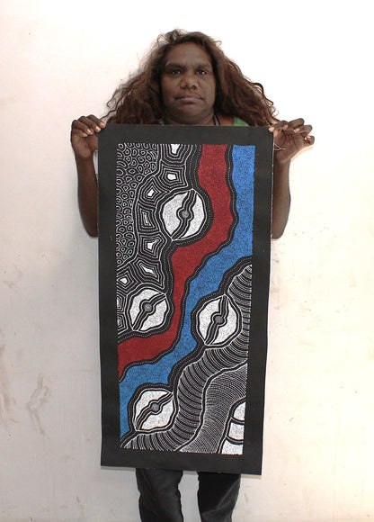 Delvine Pitjara holding her My Country painting with intricate dotted patterns in red, blue and white on black.