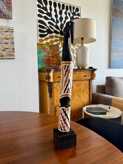 Painted milkwood bird carving by Aboriginal artist Christine Burarrwanga on a wooden table with a lamp and artwork in the background.
