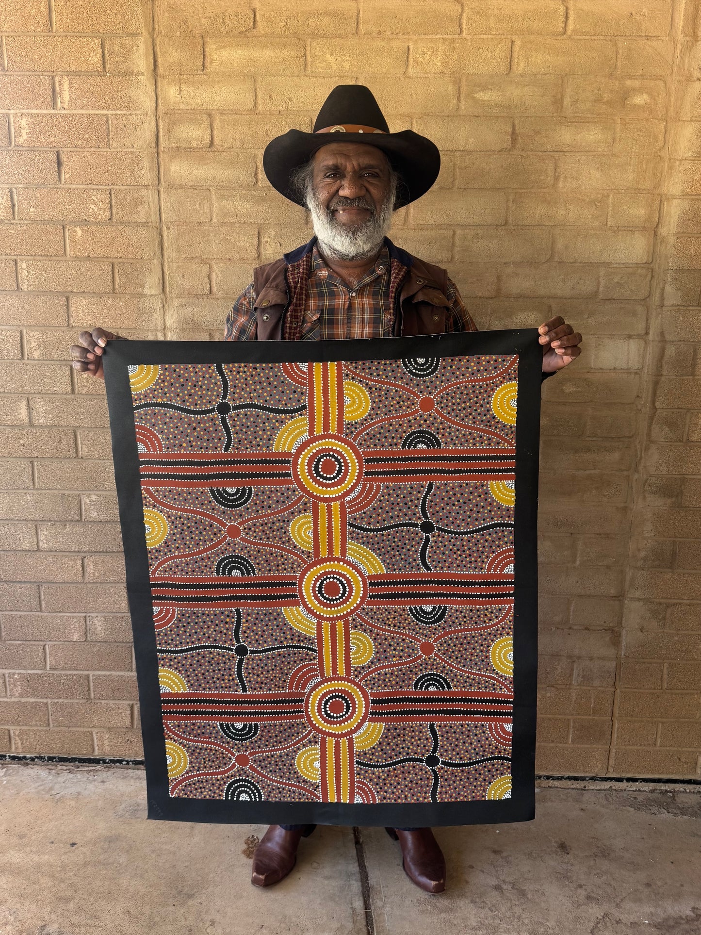 Kevin Bird Mpetyane standing against a brick wall holding his painting depicting mulga seed dreaming.