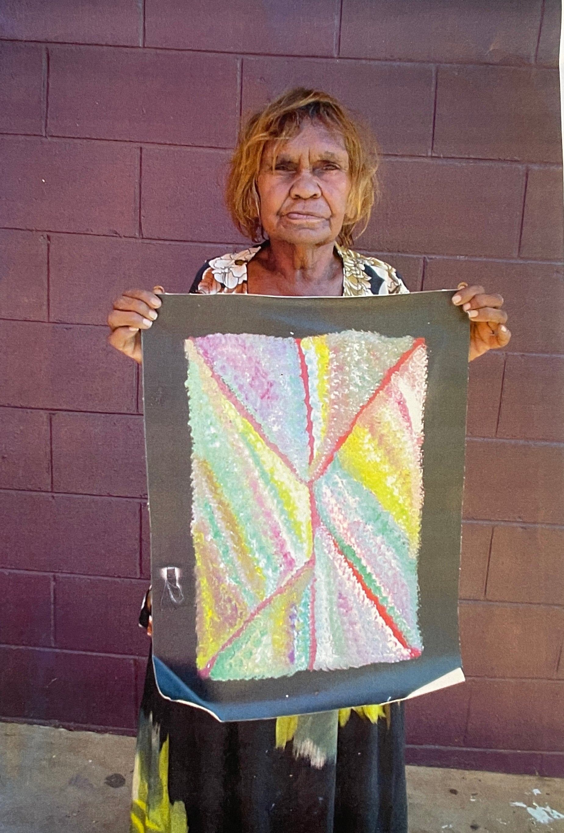 Artist Mary Morton Kemarre standing against a brick wall, holding one of her colourful paintings.