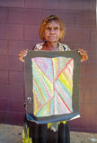 Artist Mary Morton Kemarre standing against a brick wall, holding one of her colourful paintings.