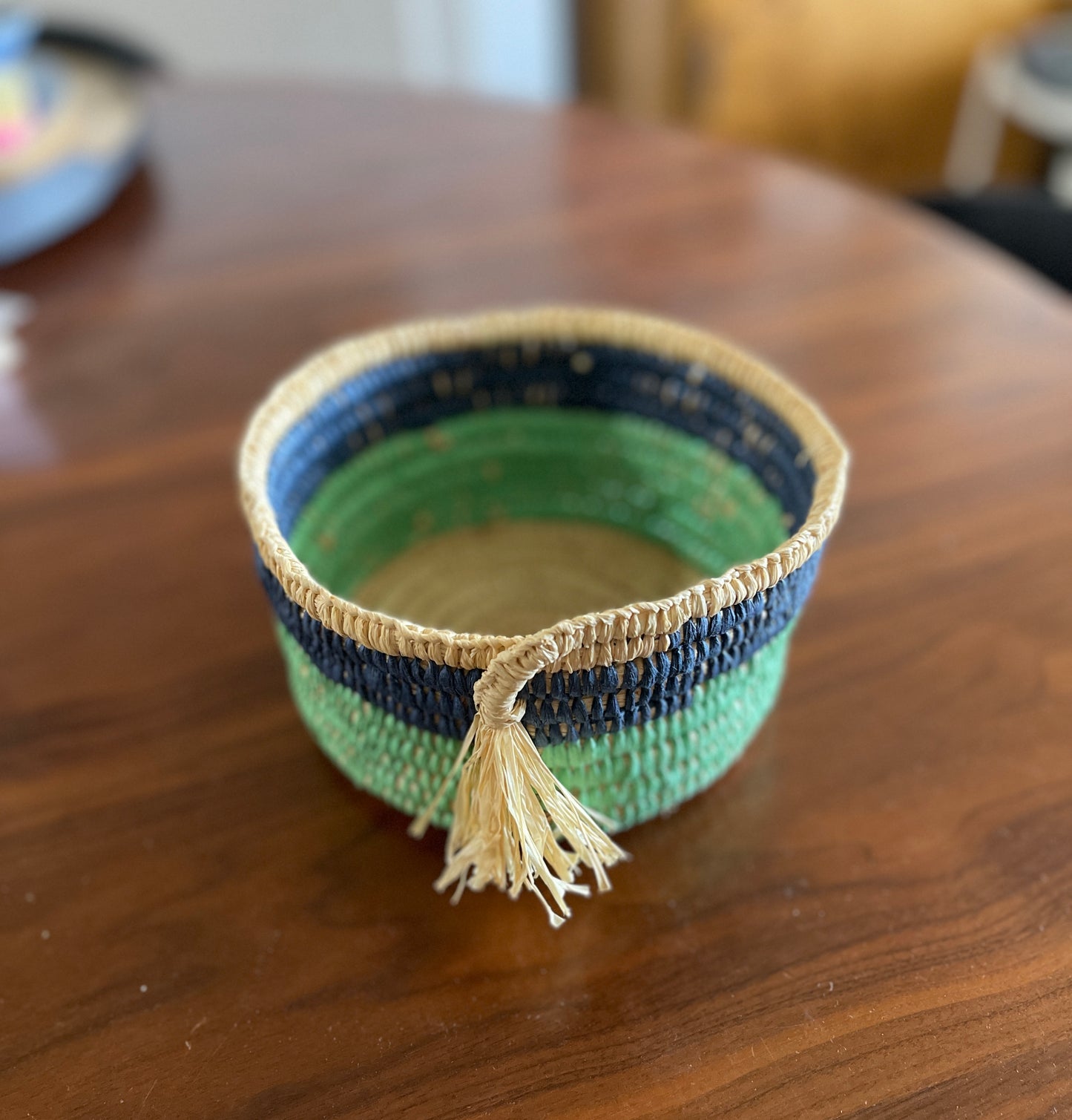 Woven basket with tassel made with green, blue and beige raffia on a wooden surface.