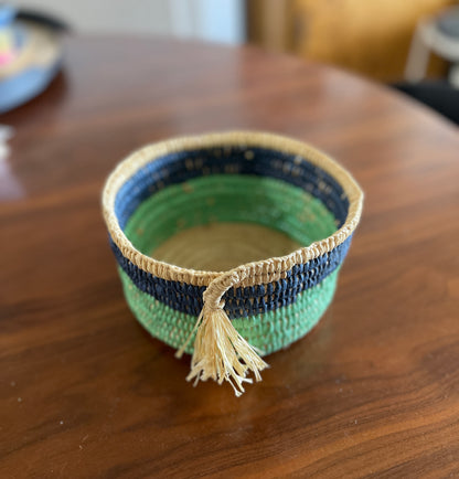 Woven basket with tassel made with green, blue and beige raffia on a wooden surface.