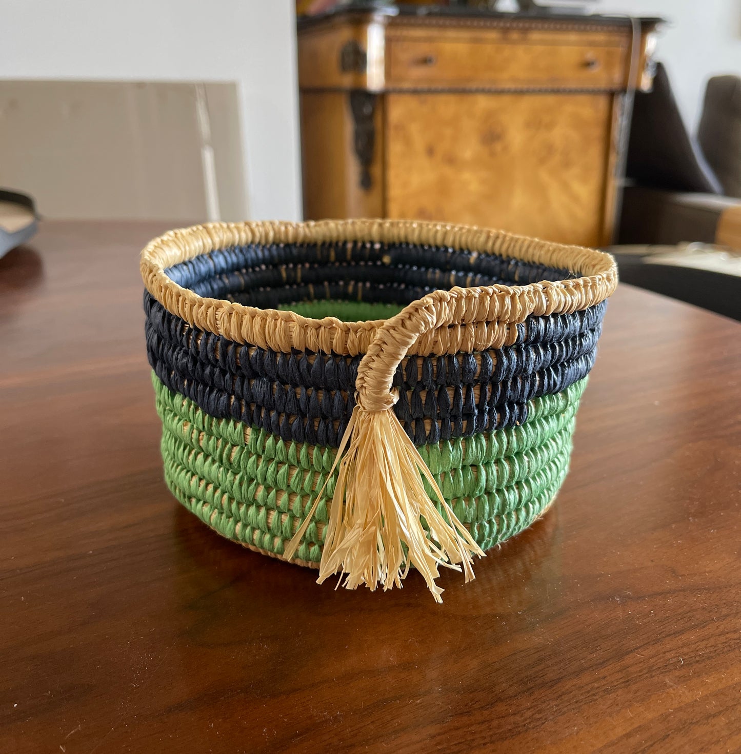 Woven basket with tassel made with green, blue and beige raffia on a wooden surface.