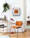 Framed artwork by Jill Daniels on a wall above an orange chair in a modern room with a gold side table and framed artwork on the wall.