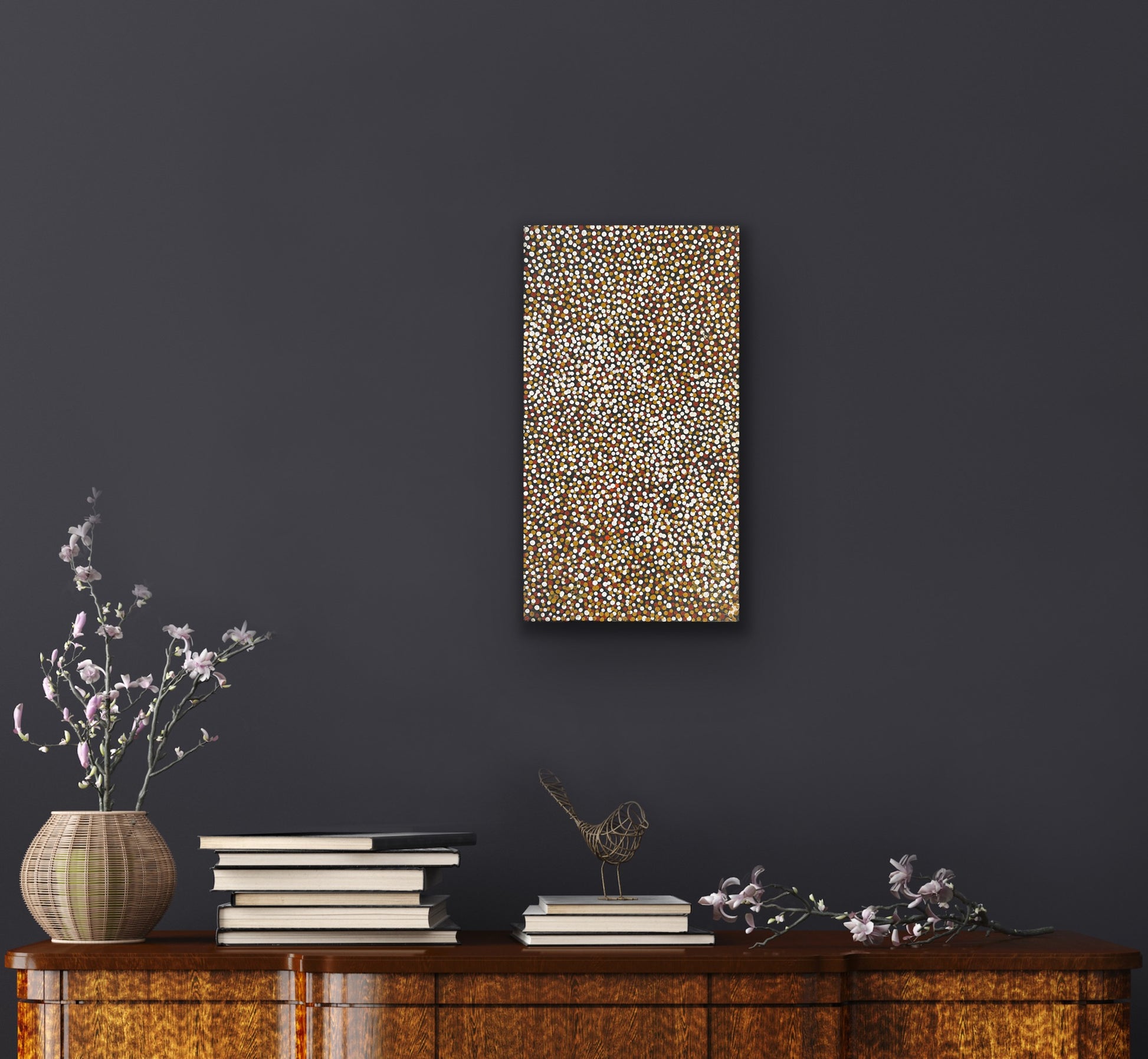 A small painting by Polly Ngale on a dark wall with a wooden cabinet below featuring books and a vase.