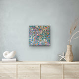 A colourful 30x30cm painting by Belinda Golder Kngwarreye on a wall above a wooden cabinet with decorative items.