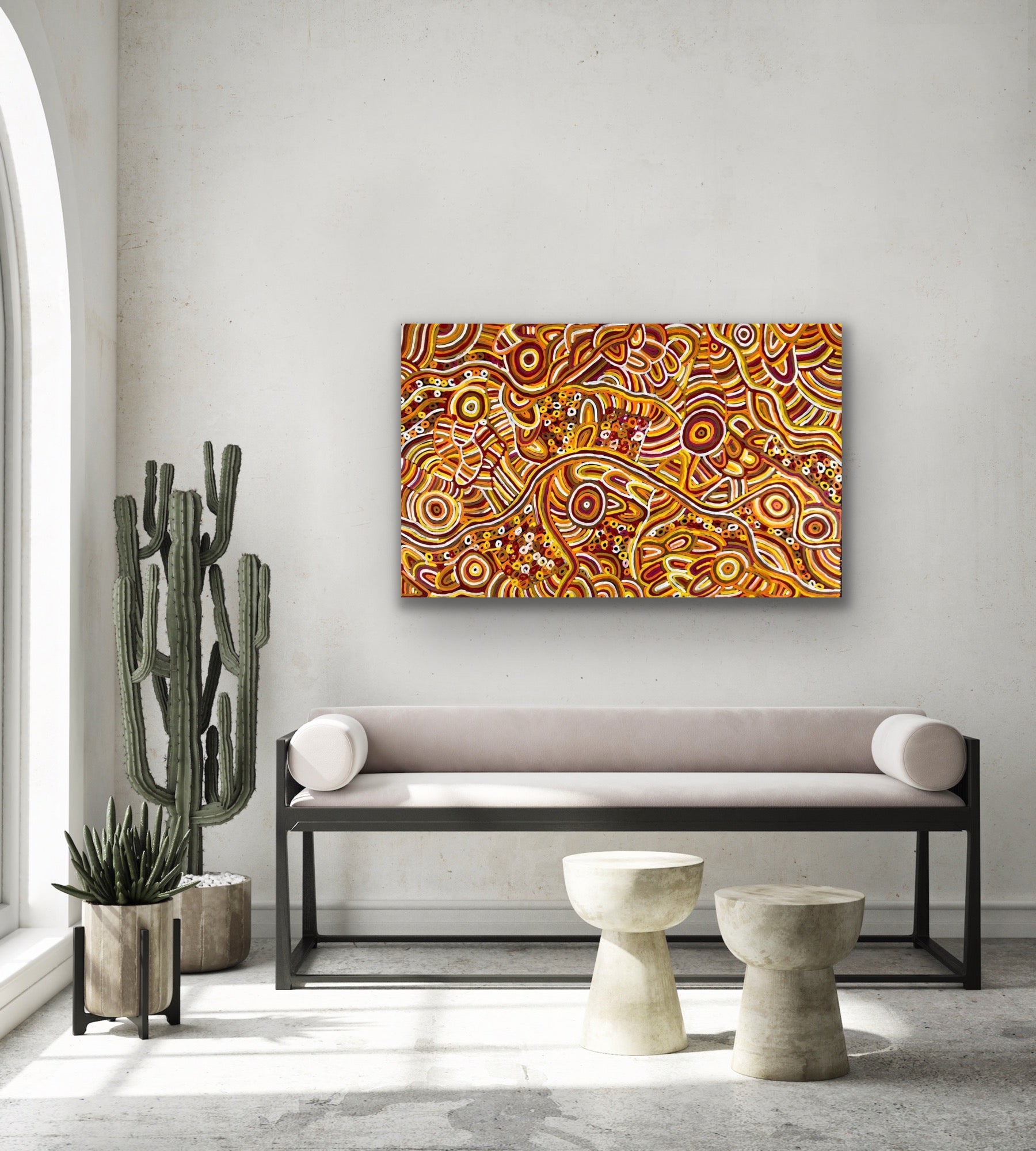 A painting by Betty Club Mbitjana hanging on a wall above a modern bench seat, plants and tables.