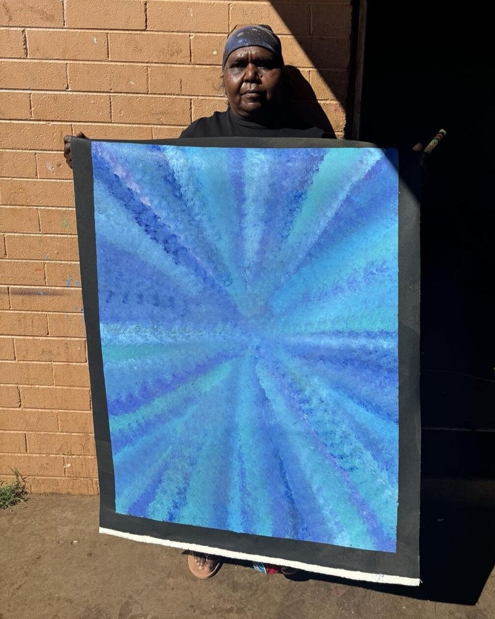 Artist Rachel Nambula standing in front for a brick wall background, holding her blue yam dreaming painting 93x71cm