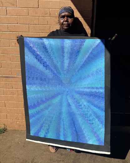Artist Rachel Nambula standing in front for a brick wall background, holding her blue yam dreaming painting 93x71cm