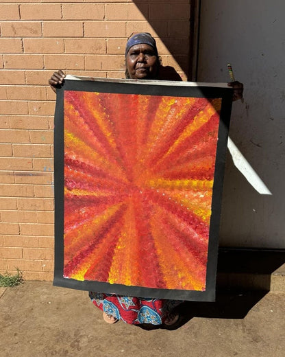 Artist Rachel Nambula standing in front for a brick wall background, holding her red and orange yam dreaming painting 93x71cm