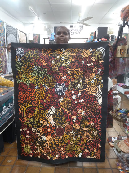 Meredith Curley holding her artwork featuring swirling patterns and a depiction of women collecting bush foods, with a predominantly brown, black, red, orange, and yellow colour palette.