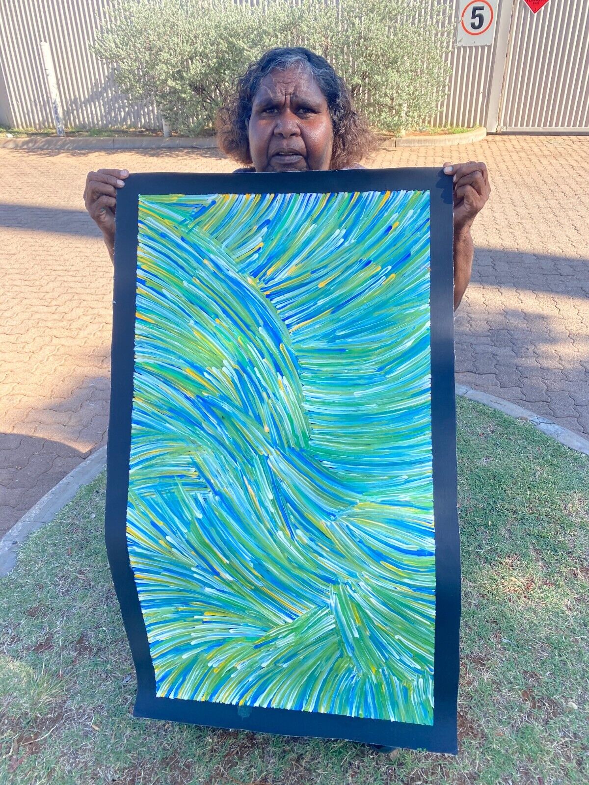 Jedda Purvis Kngwarreye holding her pencil yam painting featuring vibrant green, blue and yellow lines in flowing, traditional patterns.