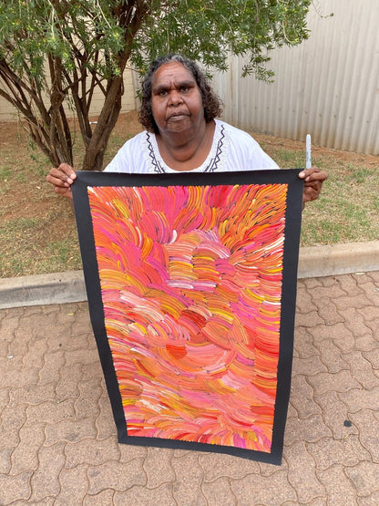 Jedda Purvis Kngwarreye holding her pencil yam painting featuring swirling patterns in pink, red, yellow and orange hues.