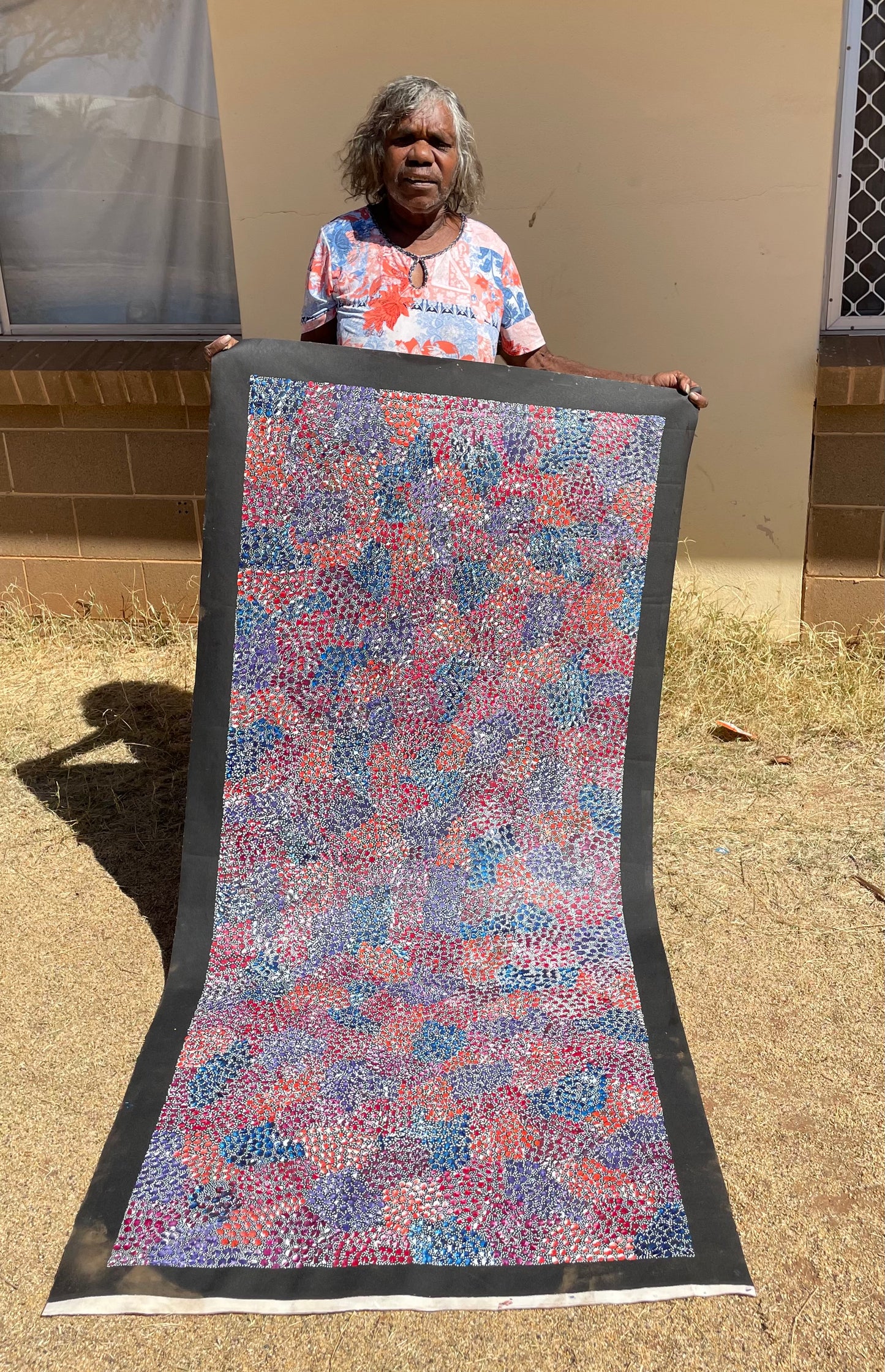 Eileen Bird Kngwarreye holding her 'Arlatyeye (Pencil Yam) Dreaming' painting 80x183cm Multi