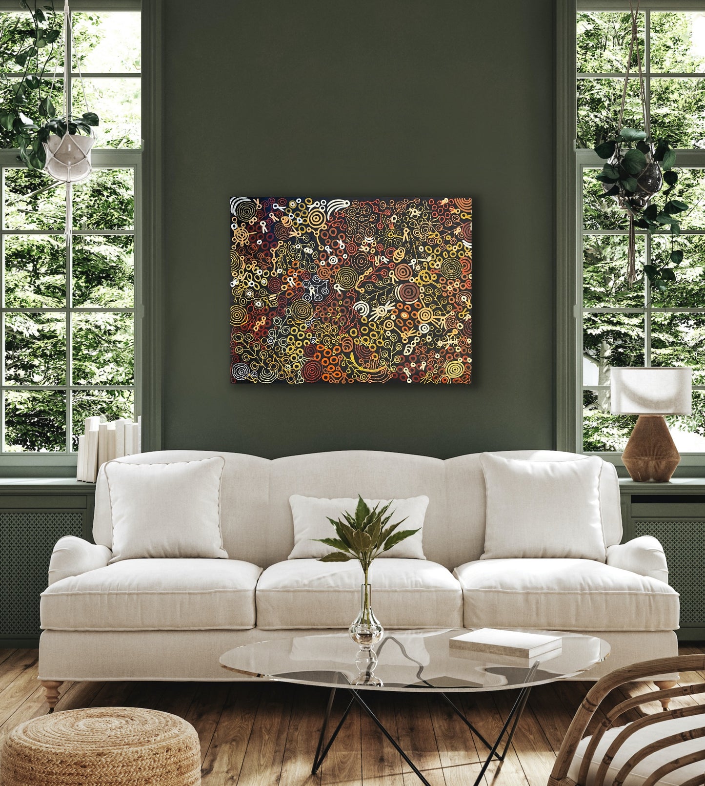 A painting by Meredith Curley hanging on a green wall above a modern white sofa, the painting features swirling patterns and symbols (a depiction of women collecting bush foods) with a predominantly brown, black, red, orange, and yellow colour palette.