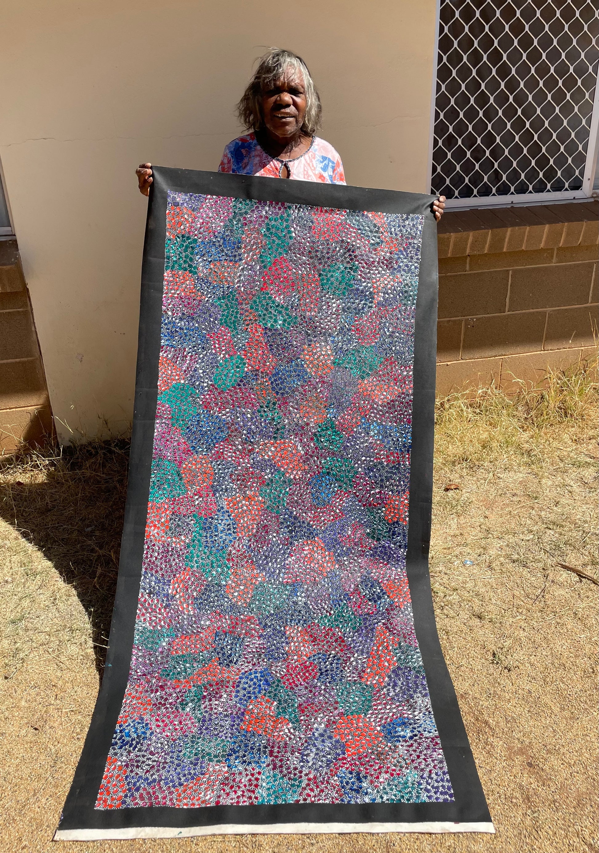 Eileen Bird Kngwarreye holding her 'Arlatyeye (Pencil Yam) Dreaming' painting 80x183cm Multi