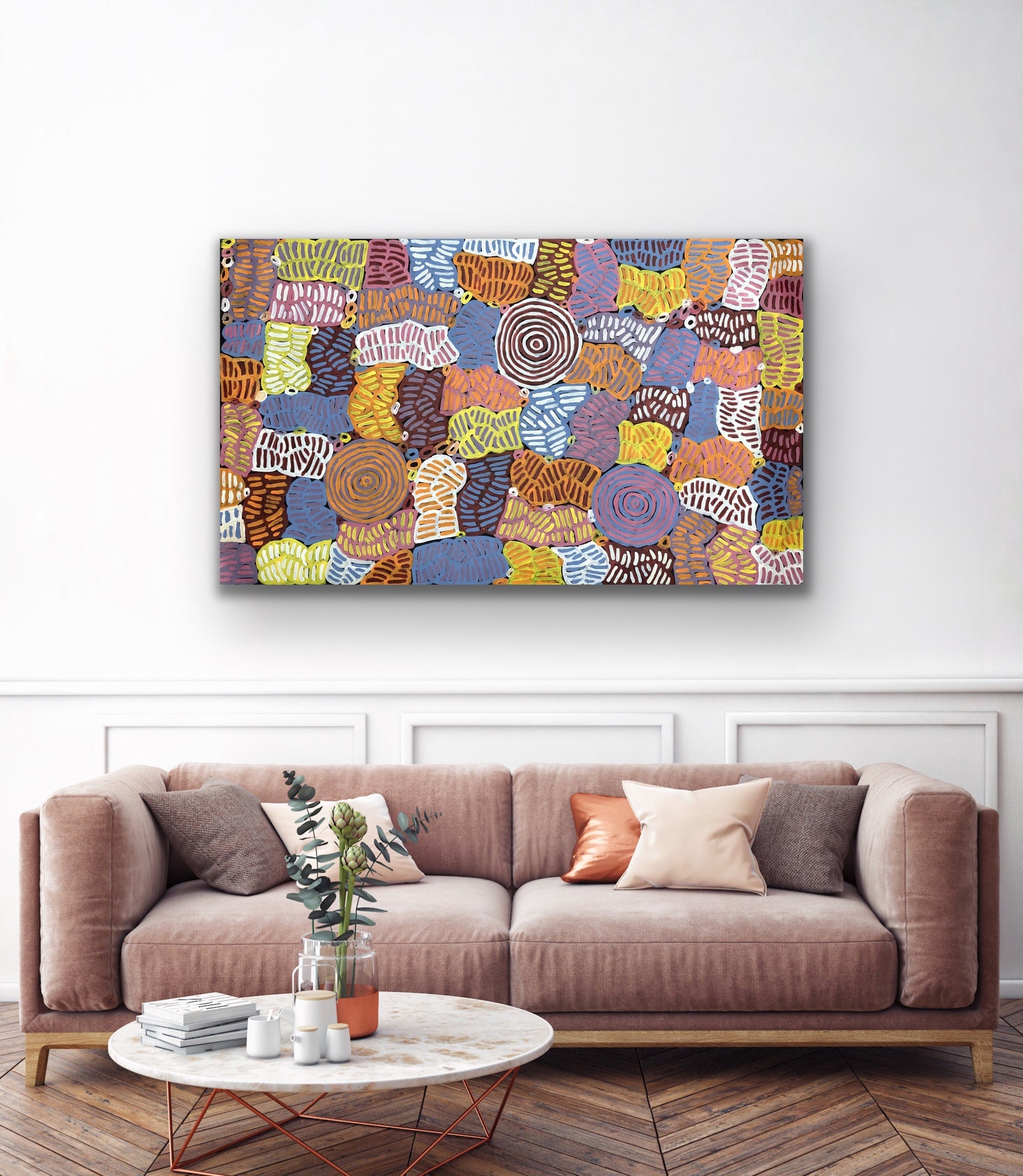 An awelye painting by Betty Club Mbitjana hanging on a white wall above a sofa and marble coffee table.