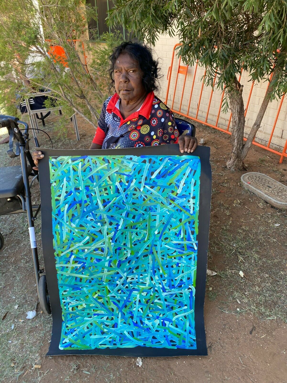 Betty Club Mbitjana holding her My Mother's Dreaming painting in predominantly blue and green hues.