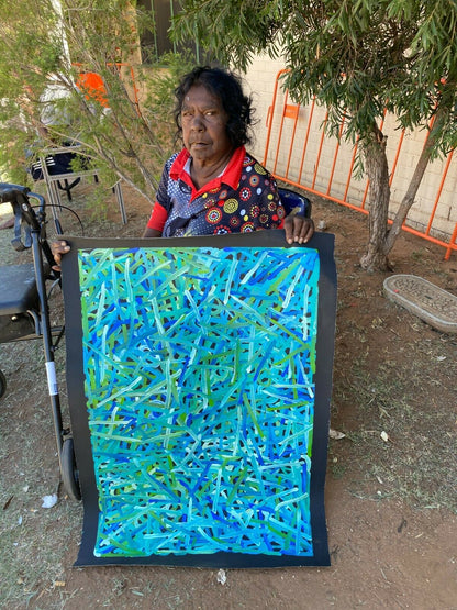 Betty Club Mbitjana holding her My Mother's Dreaming painting in predominantly blue and green hues.