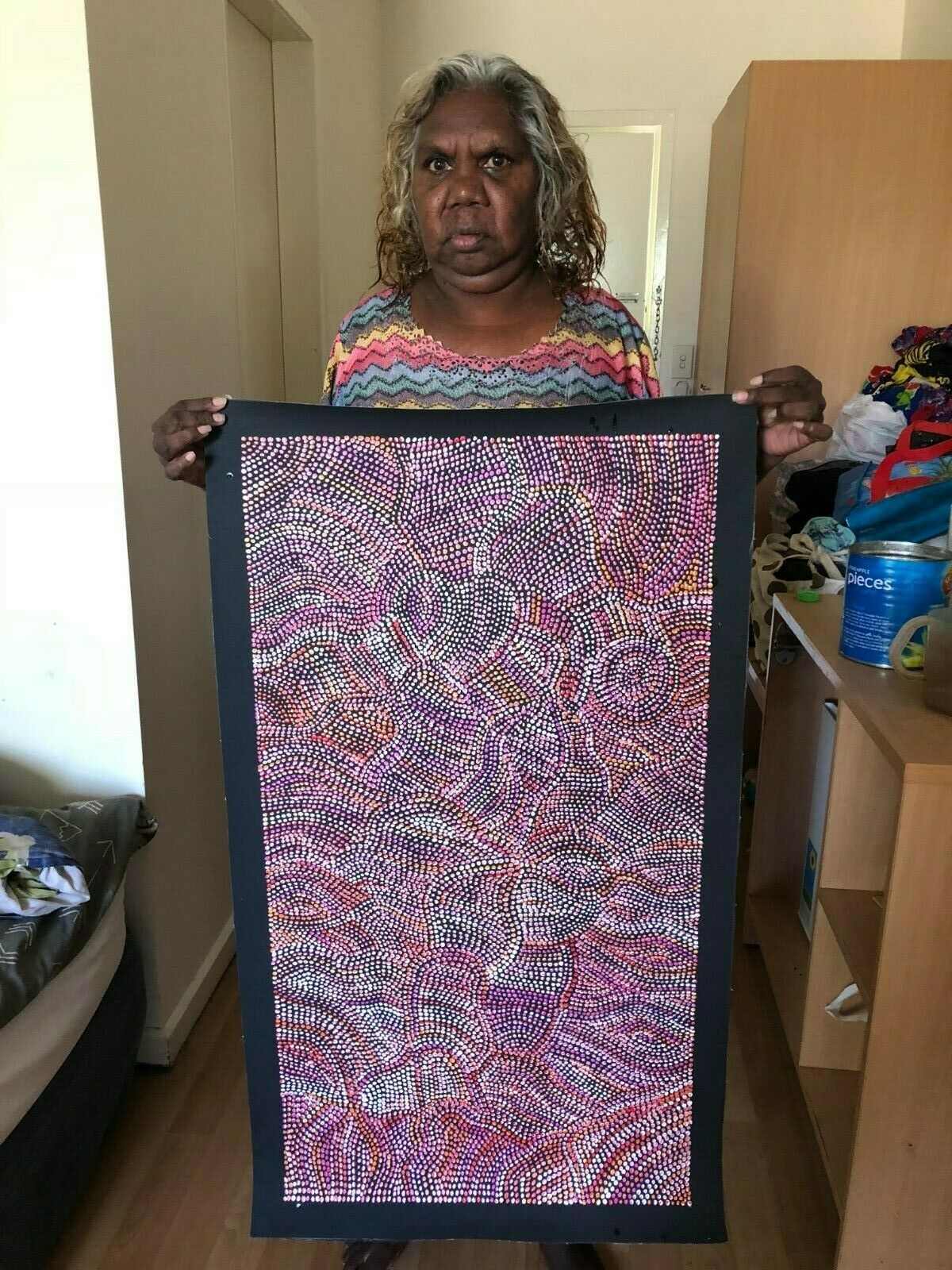 Joy Purvis Pitjara holding her Bush Yam seeds painting with a mix of red, pink, and white dots.