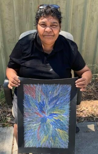 Bernadine Johnson Kemarre holding her painting with a radiant pattern in shades of blue, green, yellow, and red, representing Bush Medicine Leaves.